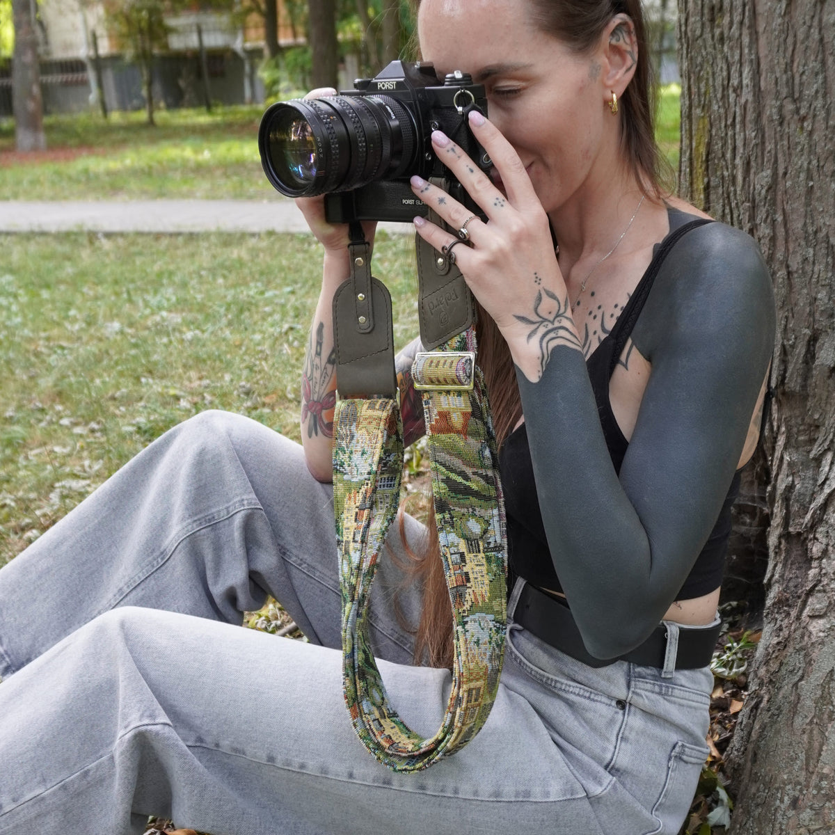 A woman sitting outdoors holds a camera with a colorful landscape-patterned camera strap. 