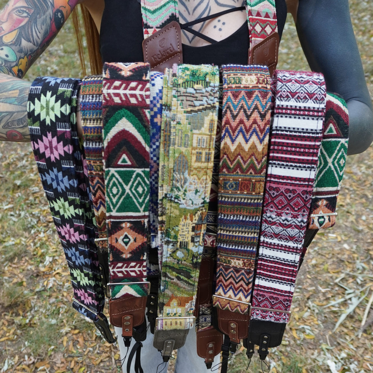 A person outdoors holding up a fan of multiple woven camera straps with colorful geometric and tribal patterns, including green, red, blue, pink, black, and brown.