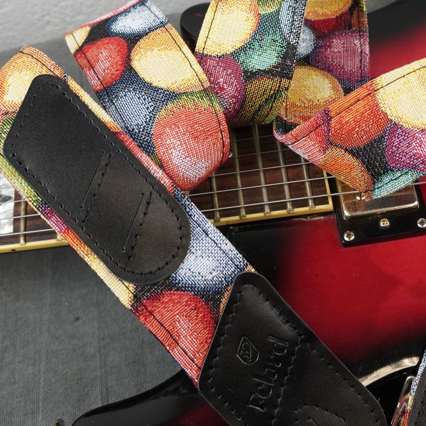 A close-up of a guitar strap with a colorful fruit print and black leather ends, shown resting on a red electric guitar.
