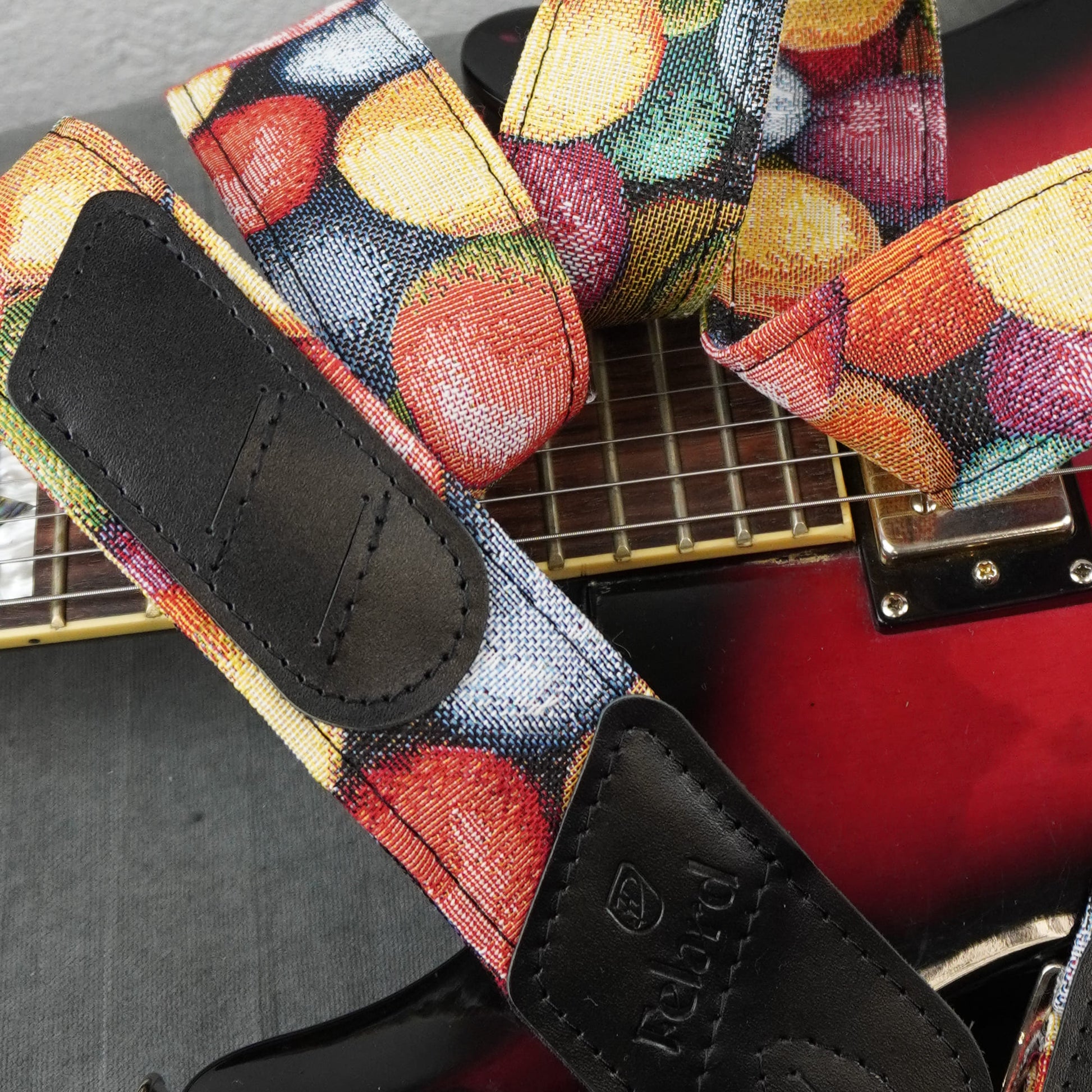 A close-up of a guitar strap with a colorful fruit print and black leather ends, shown resting on a red electric guitar.