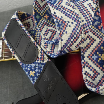 A close-up of a guitar strap with a geometric woven pattern in blue, red, yellow, and white, featuring black leather end pieces. The strap is draped over the body of a red electric guitar.