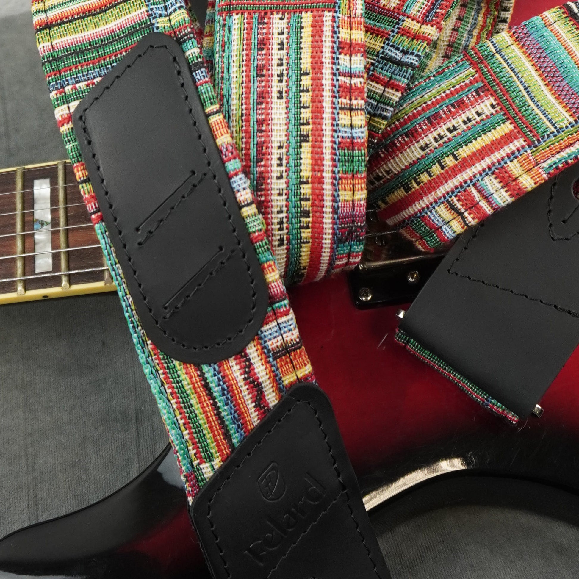 A close-up of a guitar strap with a vibrant multicolor woven pattern and black leather end pieces, shown attached to an electric guitar.