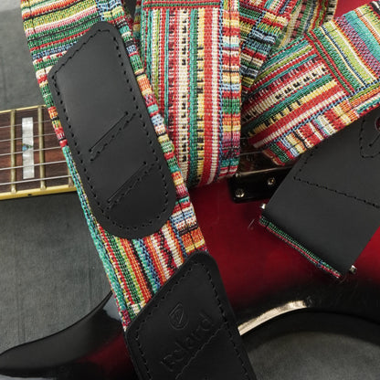 A close-up of a guitar strap with a vibrant multicolor woven pattern and black leather end pieces, shown attached to an electric guitar.