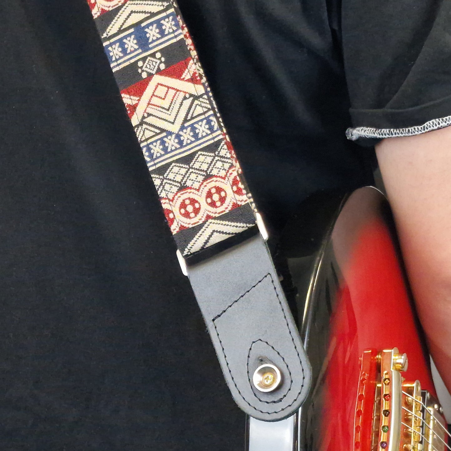 A close-up of a guitar strap worn by a person holding a red electric guitar. The strap features a geometric tribal pattern in red, beige, and navy blue, with a dark gray leather end piece and a gold-tone metal snap button.