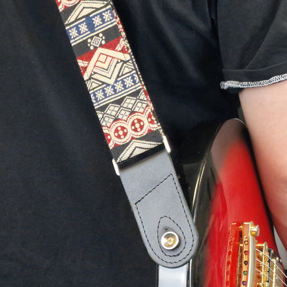 A close-up of a guitar strap worn by a person holding a red electric guitar. The strap features a geometric tribal pattern in red, beige, and navy blue, with a dark gray leather end piece and a gold-tone metal snap button.