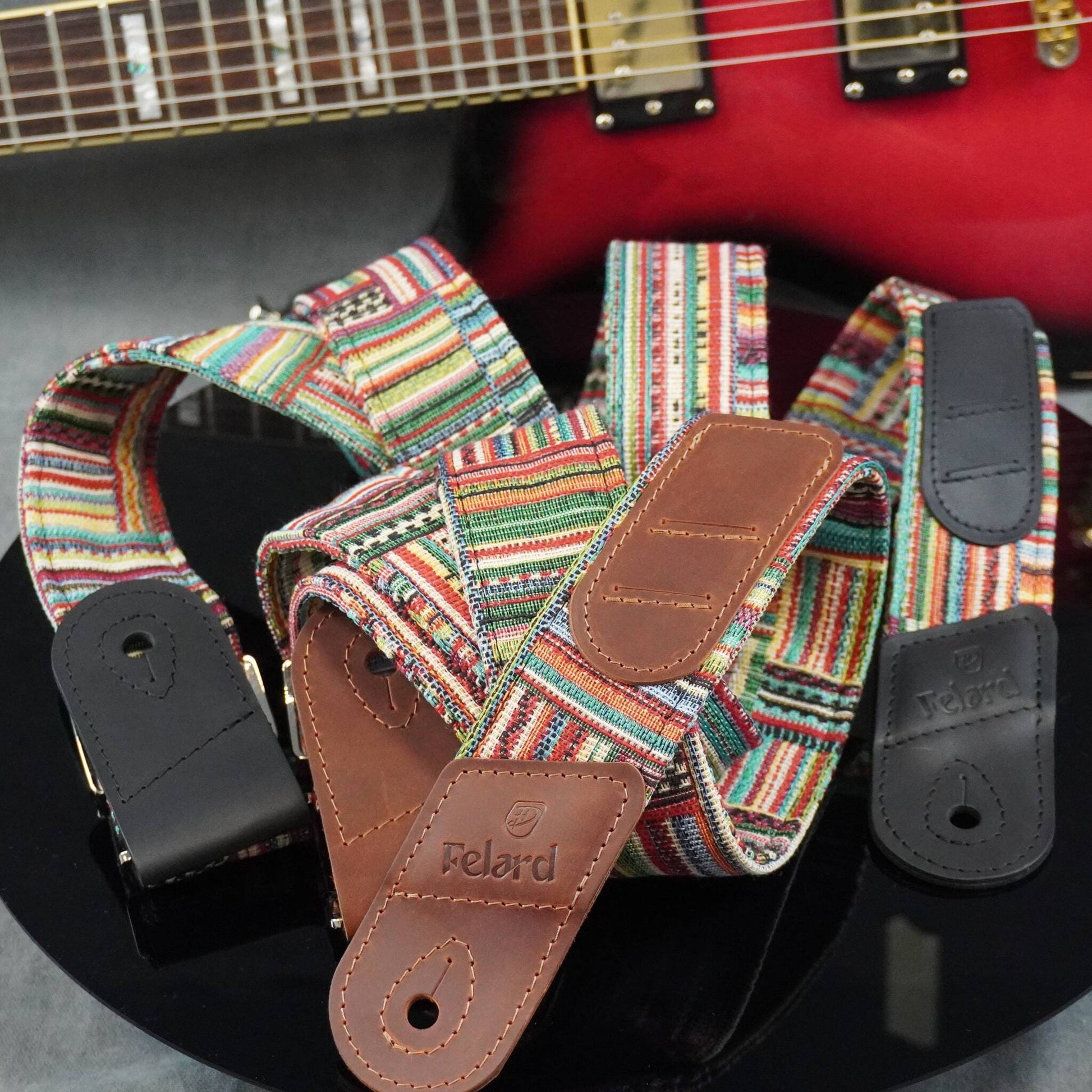 guitar strap with a colorful pattern and brown leather ends, with the brand name 'Feldrd' visible on the leather. A red electric guitar is partially visible in the background.