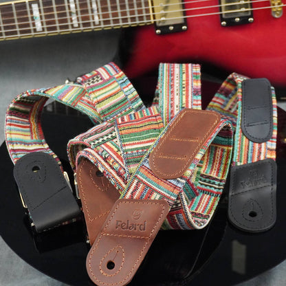 guitar strap with a colorful pattern and brown leather ends, with the brand name 'Feldrd' visible on the leather. A red electric guitar is partially visible in the background.