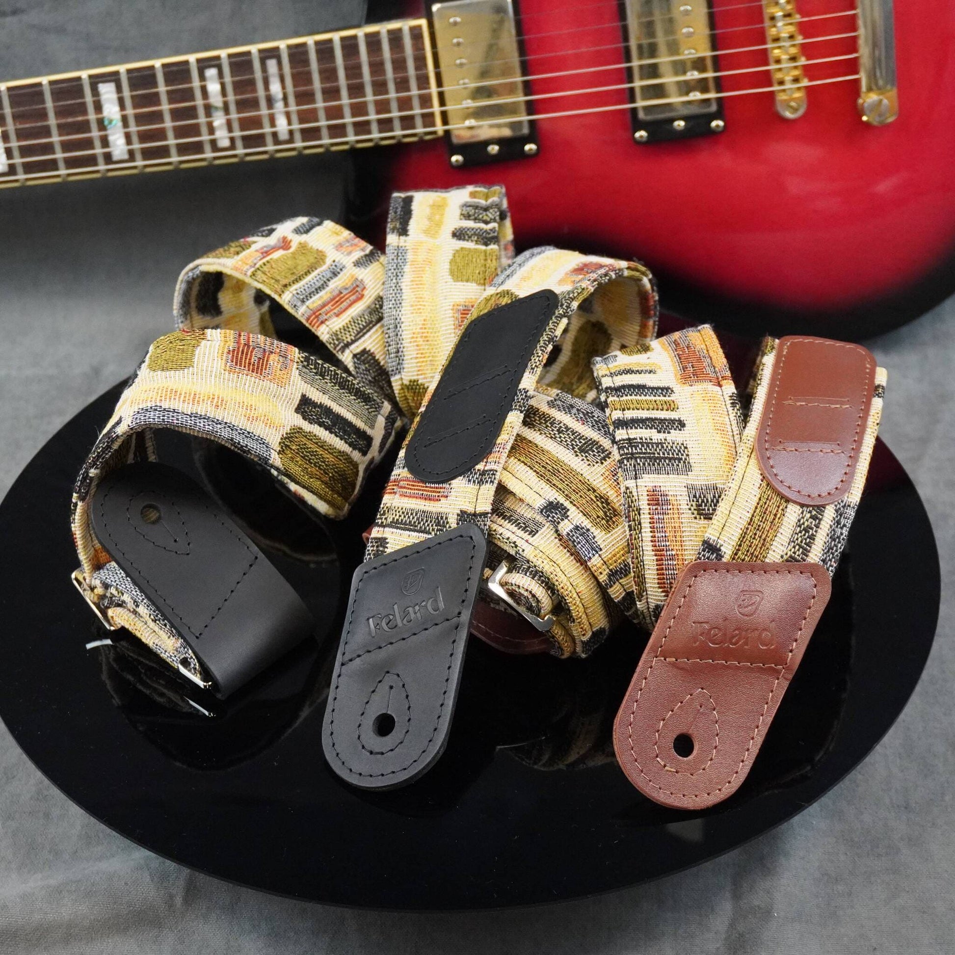 guitar strap cases with patterned fabric and leather ends displayed on a black plate, with a red electric guitar blurred in the background.