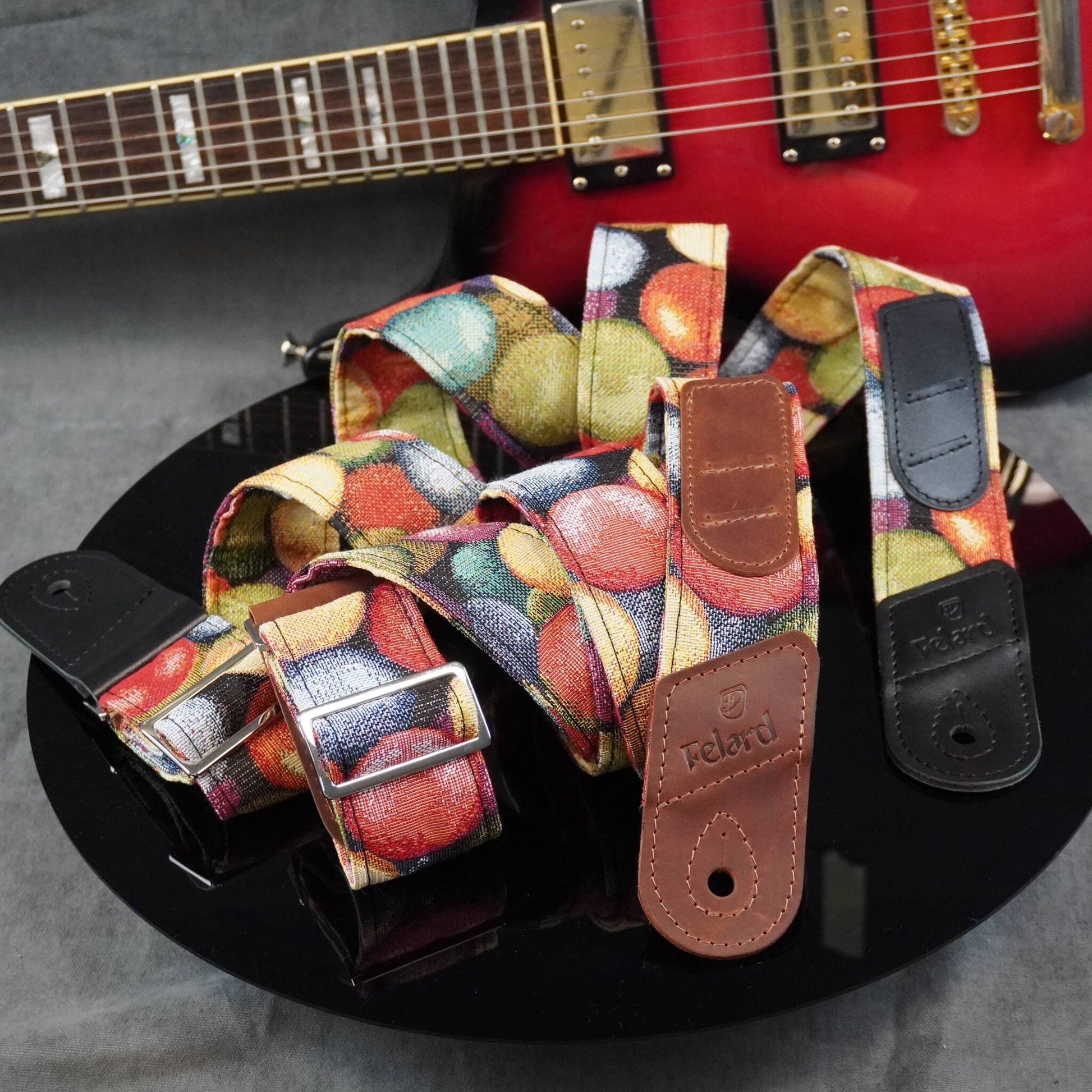 Colorful guitar strap with a multicolor fruit print and brown leather ends, displayed on a black reflective surface with a red electric guitar in the background.