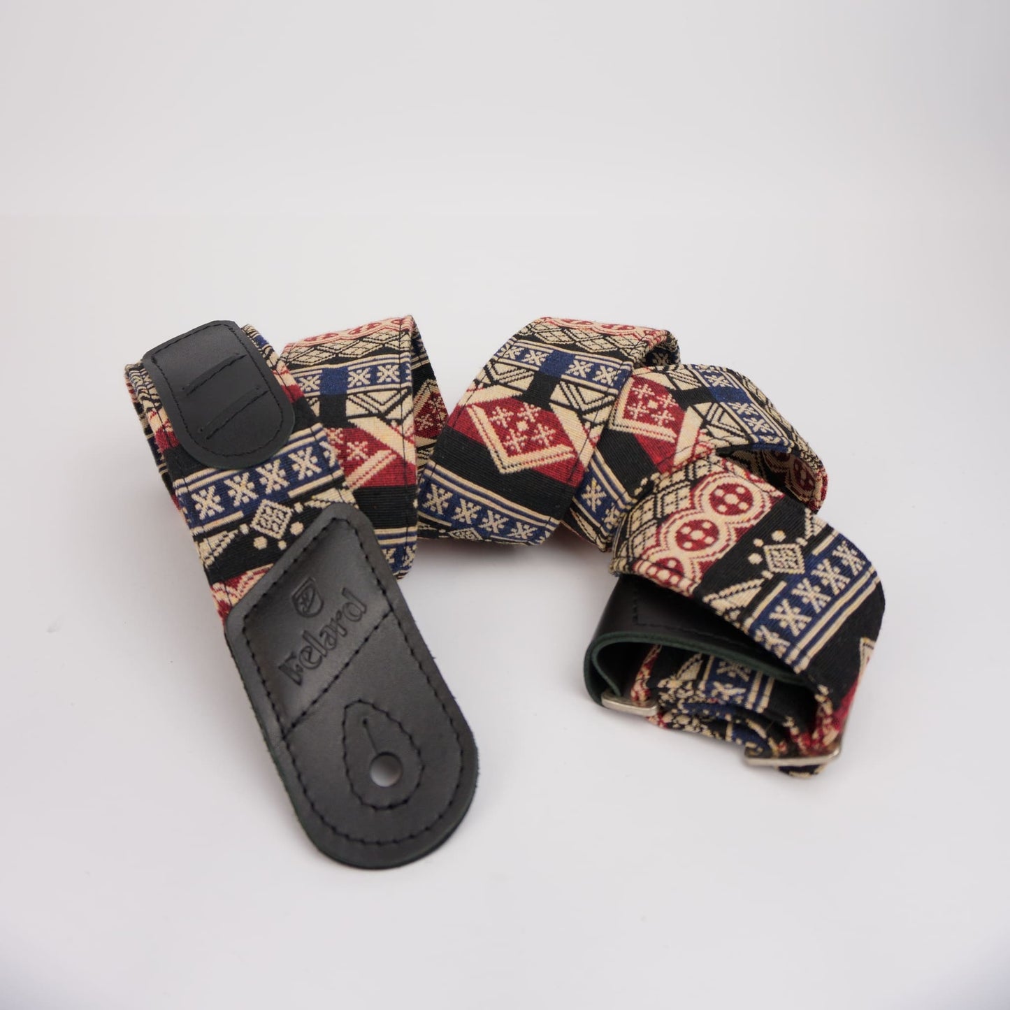 patterned guitar strap with a black leather  and a geometric print featuring red, blue, beige, and black, displayed on a light gray background.