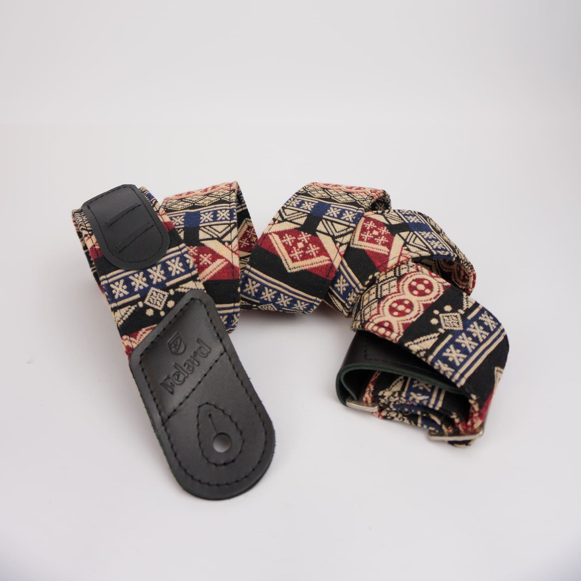 patterned guitar strap with a black leather  and a geometric print featuring red, blue, beige, and black, displayed on a light gray background.