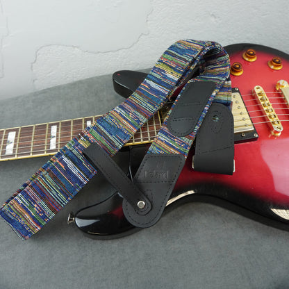 A close-up of an electric guitar strap with a multicolor woven pattern and black leather accents, draped over a red electric guitar on a gray surface.