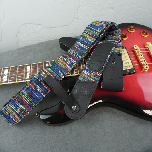 A close-up of an electric guitar strap with a multicolor woven pattern and black leather accents, draped over a red electric guitar on a gray surface.