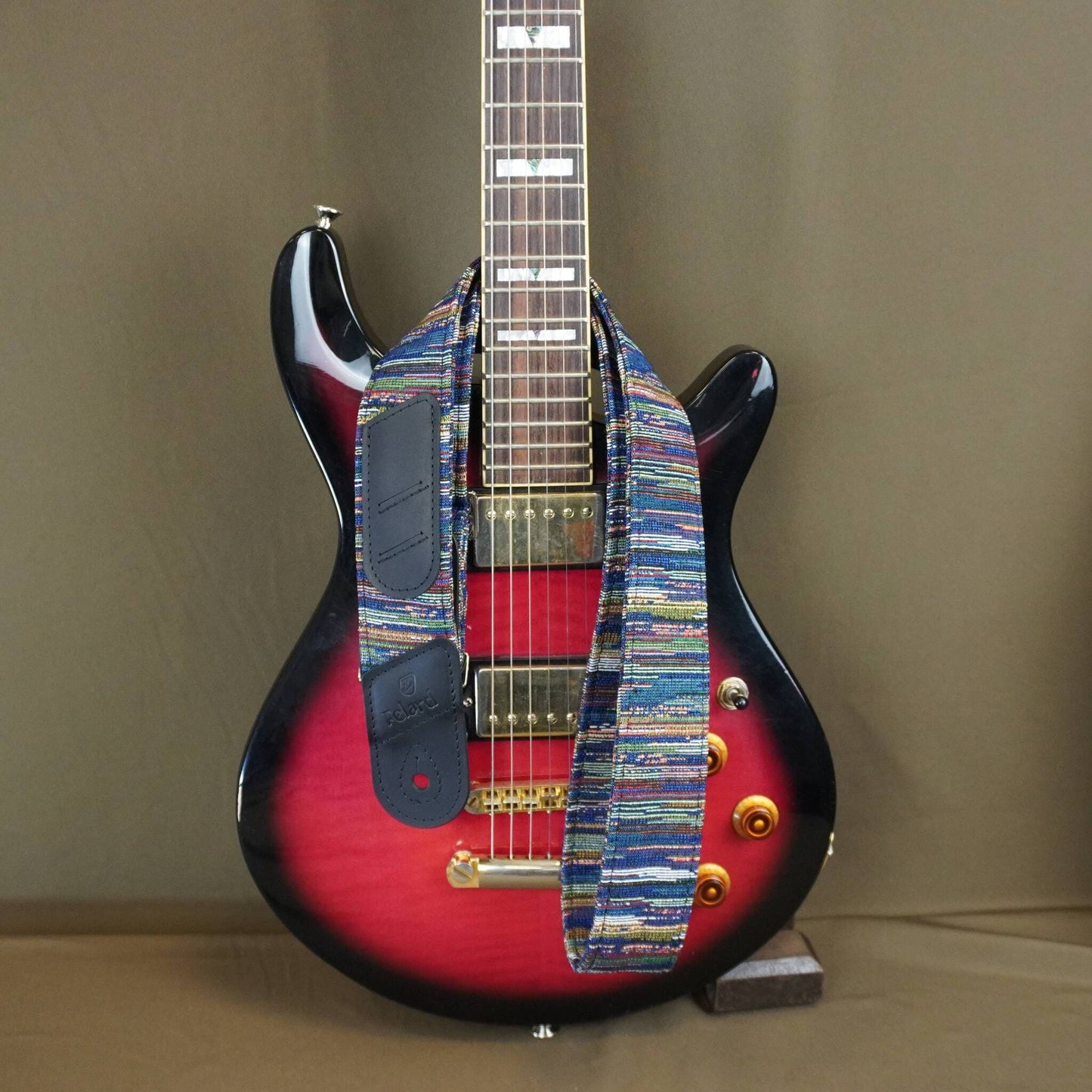 A custom electric guitar with a red and black sunburst body, featuring a colorful woven strap across the pickups and neck, set against a plain brown background.