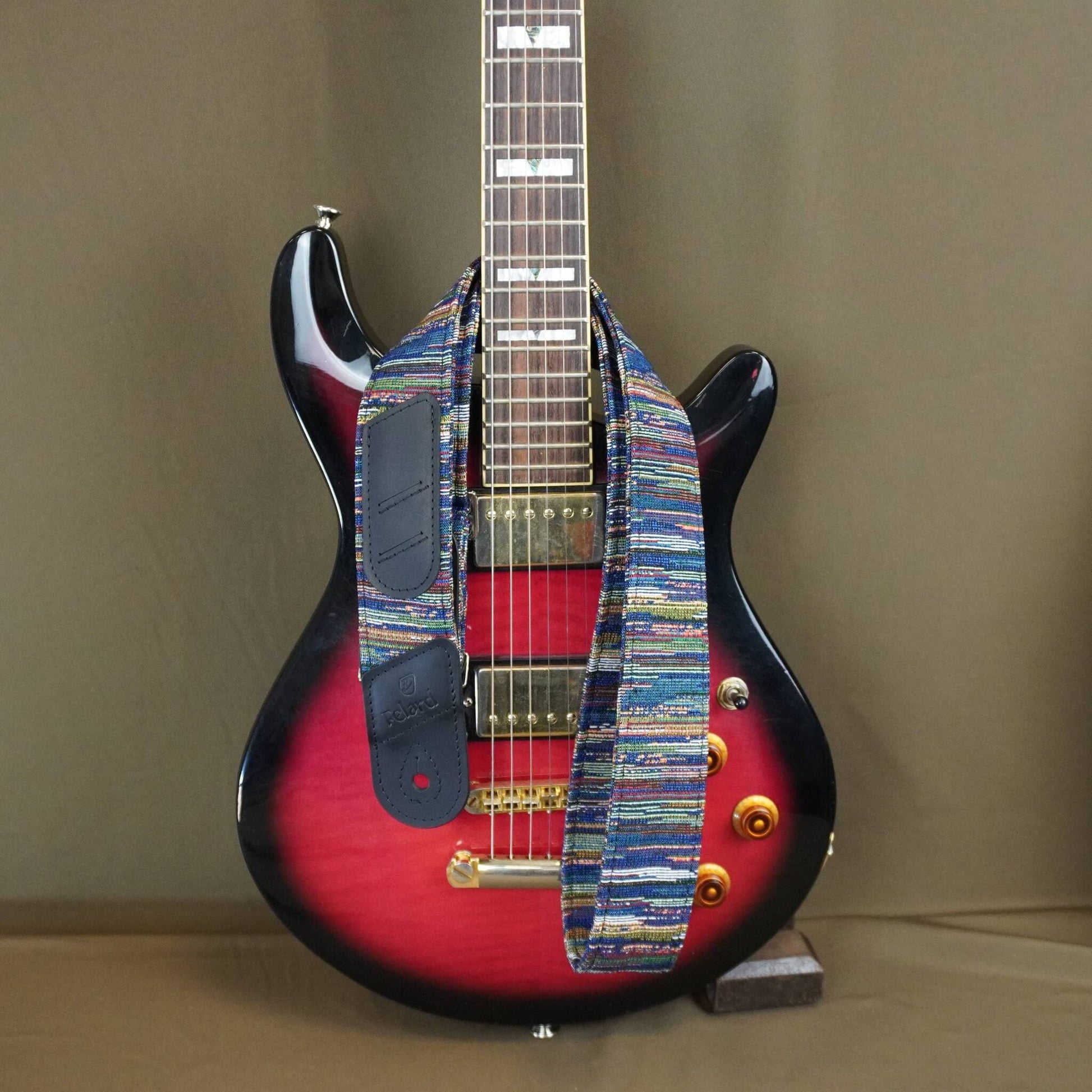 A custom electric guitar with a red and black sunburst body, featuring a colorful woven strap across the pickups and neck, set against a plain brown background.