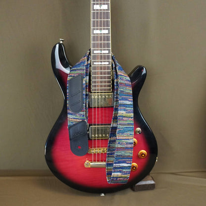 A custom electric guitar with a red and black sunburst body, featuring a colorful woven strap across the pickups and neck, set against a plain brown background.