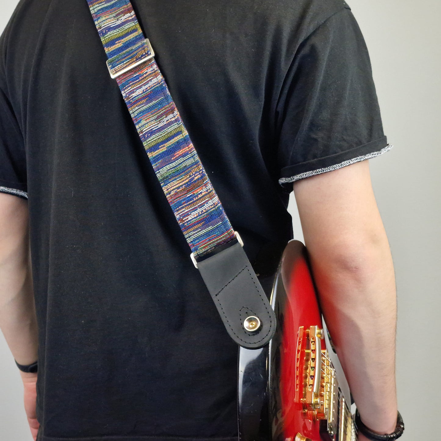 A close-up of a person carrying a red electric guitar, showing a colorful striped guitar strap with a black leather end piece.