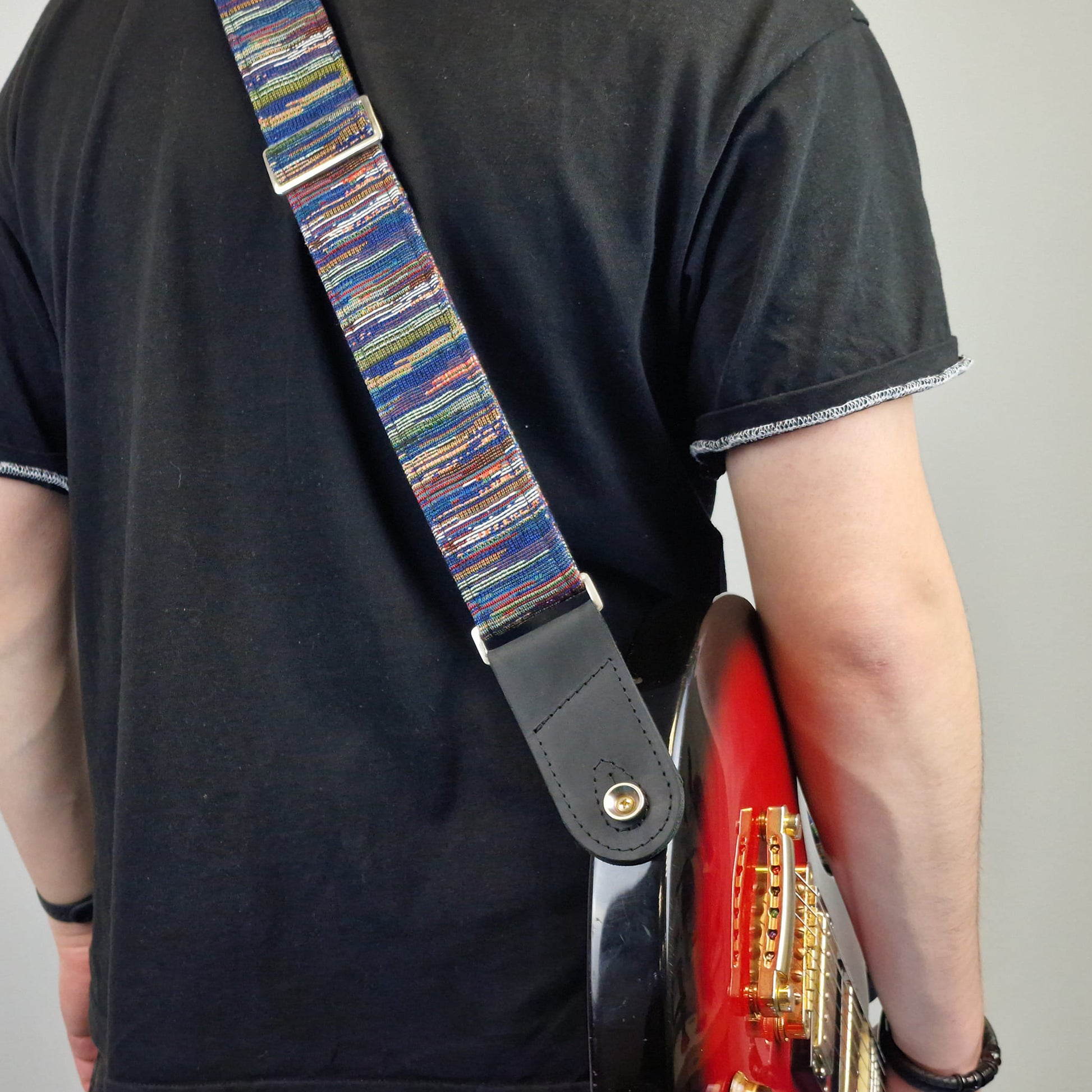 A close-up of a person carrying a red electric guitar, showing a colorful striped guitar strap with a black leather end piece.