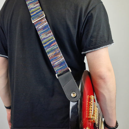A close-up of a person carrying a red electric guitar, showing a colorful striped guitar strap with a black leather end piece.