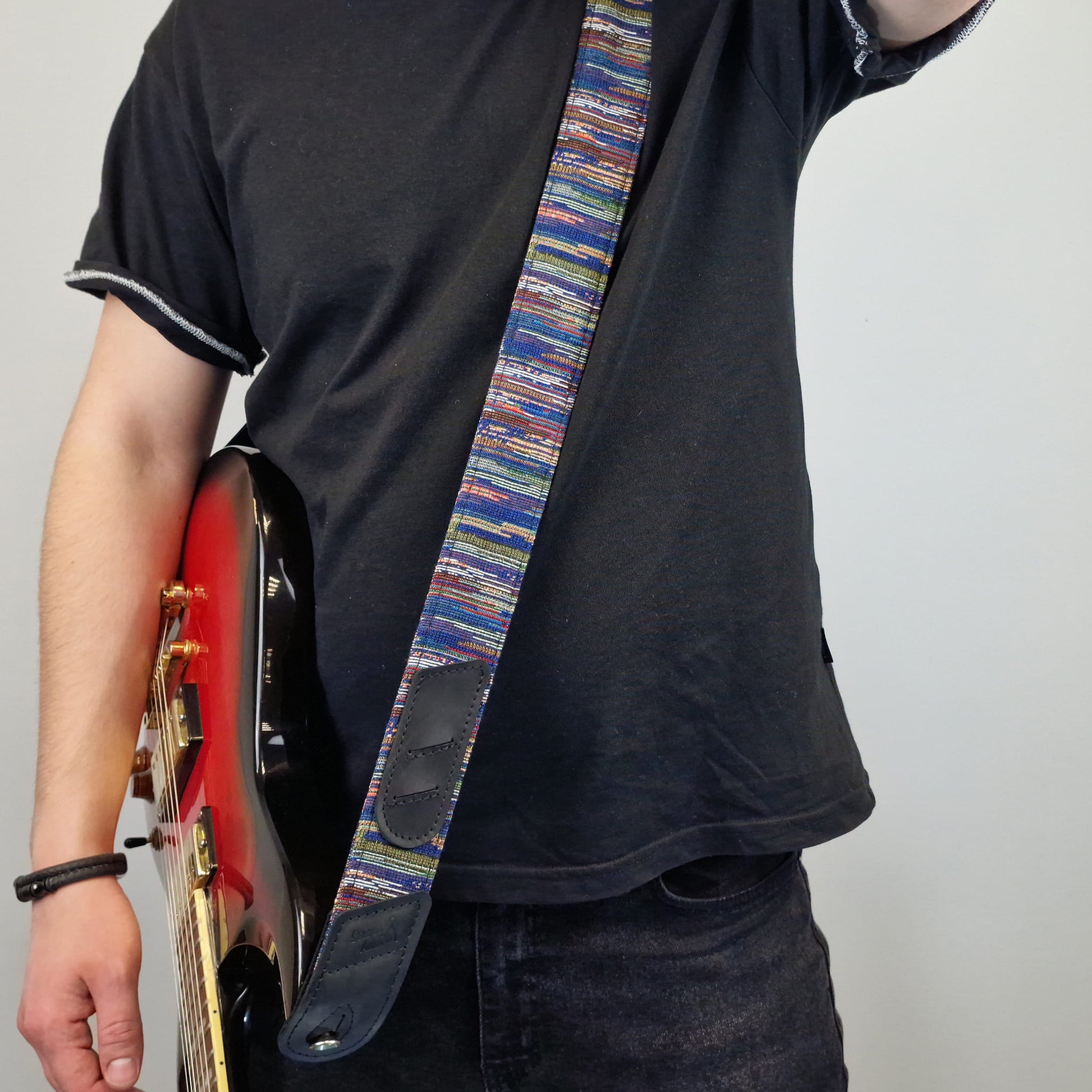 A person  carries a red electric guitar while wearing a colorful striped guitar strap with dark leather patches. The background is a plain light gray wall.