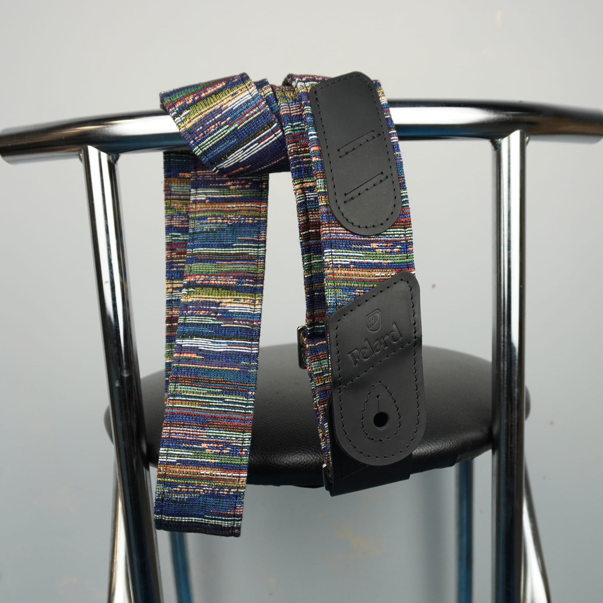 A multicolor striped fabric core guitar strap with black leather end pieces, draped over the back of a metal chair.
