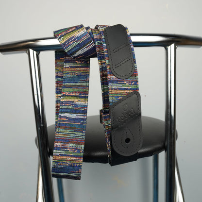 A multicolor striped fabric core guitar strap with black leather end pieces, draped over the back of a metal chair.