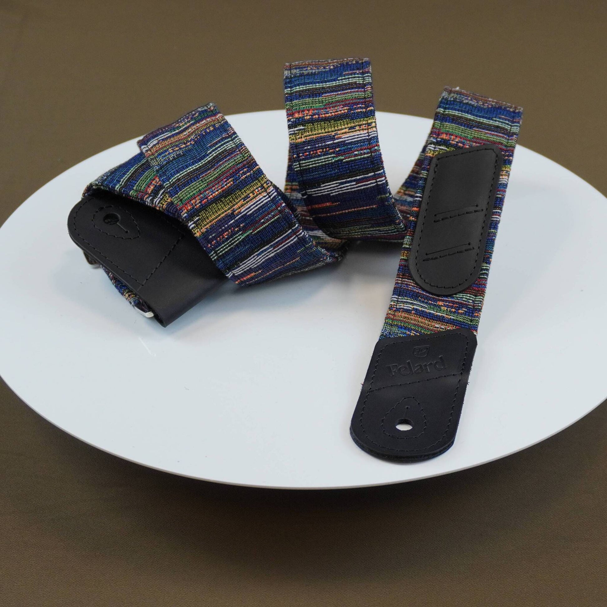 A guitar strap with a multicolor striped fabric body and black leather ends, displayed on a white plate against a brown background.