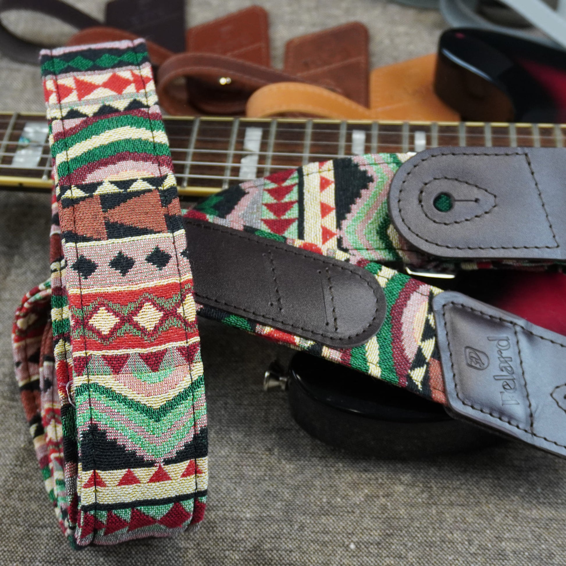 A close-up of a guitar strap with a colorful geometric Southwestern pattern in red, green, beige, and black, featuring dark gray leather accents and a visible 'Pembert' brand logo on the leather end.