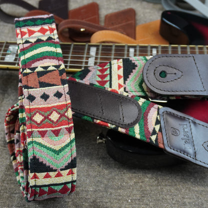 A close-up of a guitar strap with a colorful geometric Southwestern pattern in red, green, beige, and black, featuring dark gray leather accents and a visible 'Pembert' brand logo on the leather end.
