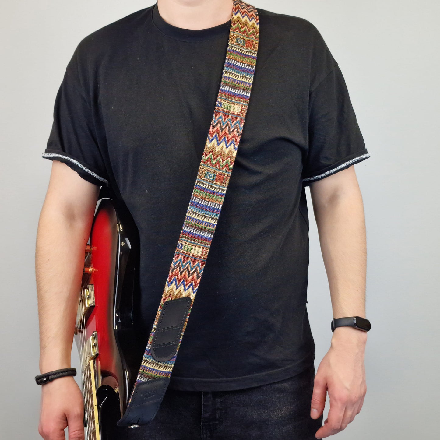 holding a red electric guitar and wearing a colorful patterned guitar strap with a geometric design.