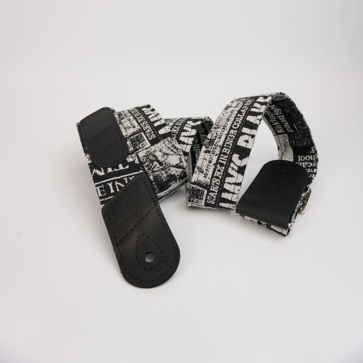 A guitar strap with a black and white newspaper print pattern featuring the text 'Felard', finished with light black leather ends and a silver metal buckle, displayed on a clean white background.