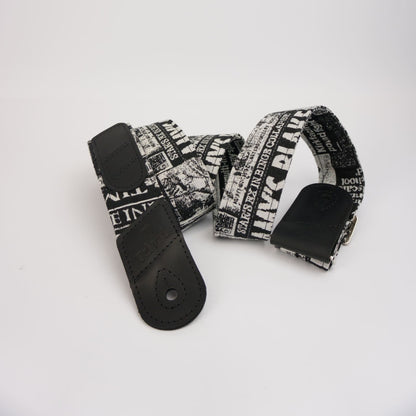 A guitar strap with a black and white newspaper print pattern featuring the text 'Felard', finished with light black leather ends and a silver metal buckle, displayed on a clean white background.