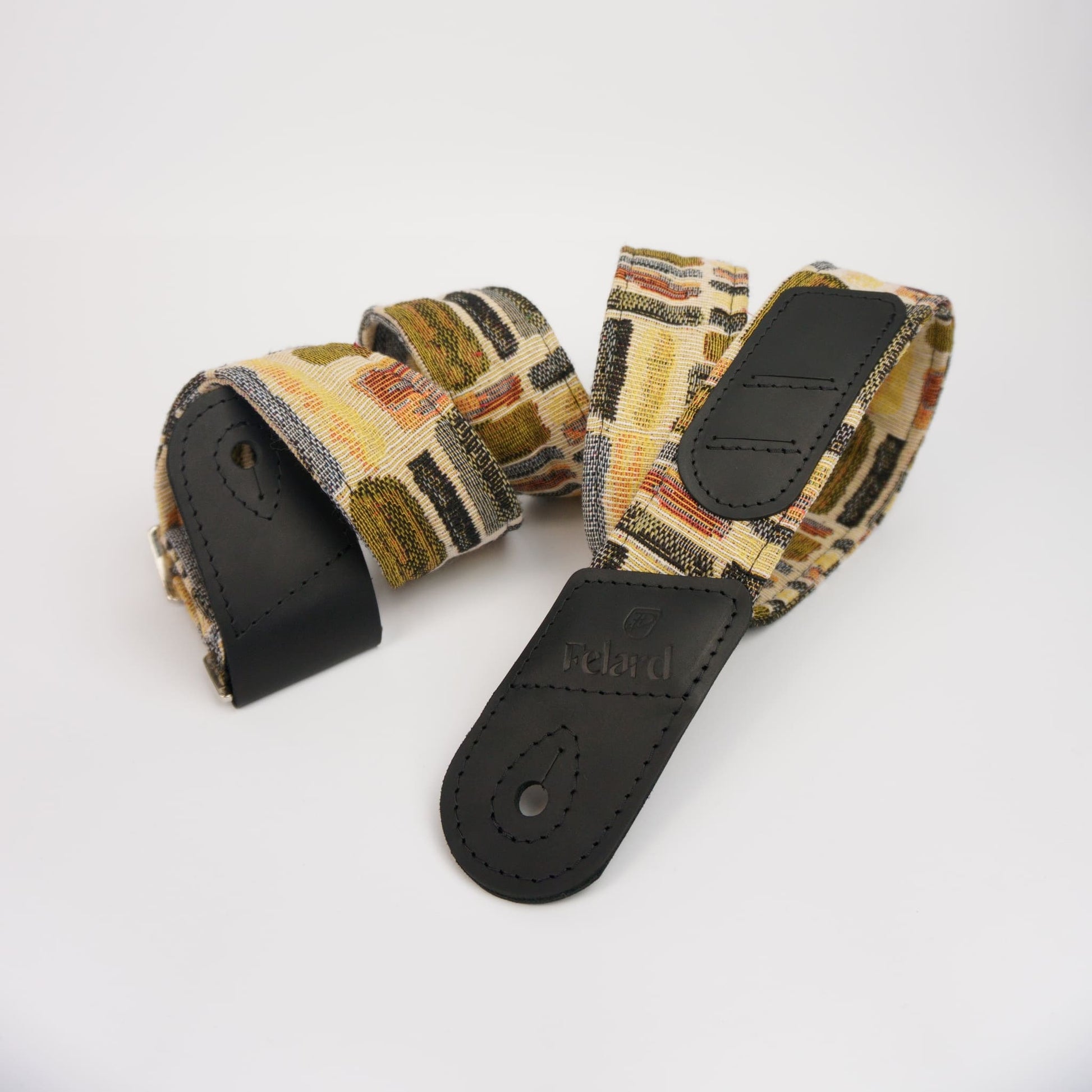 Guitar strap with a multicolor geometric pattern in beige, brown, and black, featuring black leather end caps and a buckle closure.