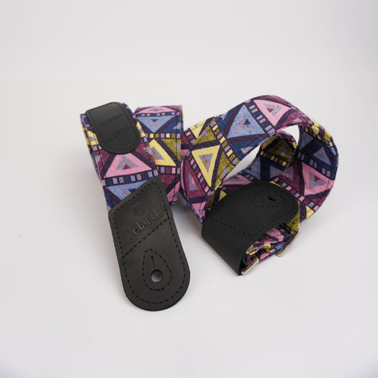 Guitar strap with a colorful geometric pattern in purple, pink, blue, and yellow, featuring black leather end caps, photographed on a plain white background.