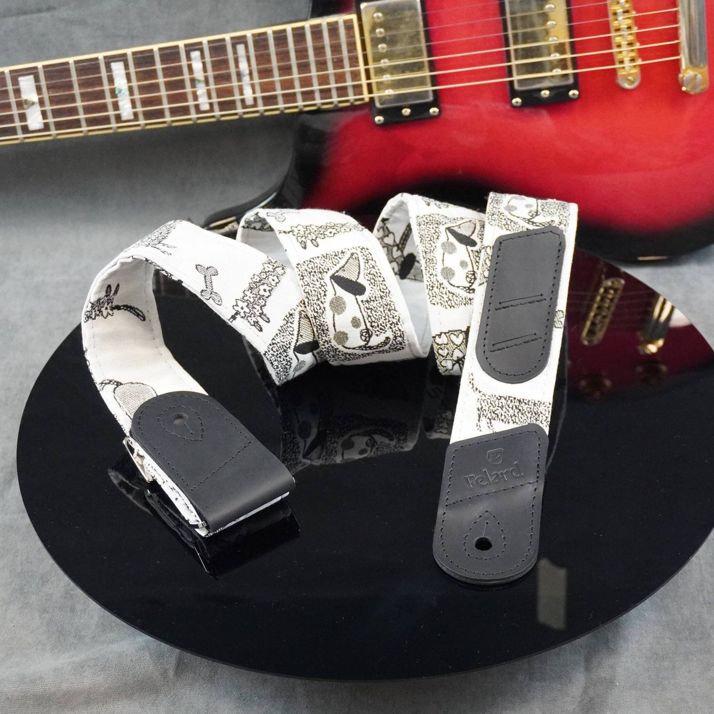 A white electric guitar strap with a black paisley pattern and black leather ends, displayed on a black reflective surface with a red electric guitar in the background.