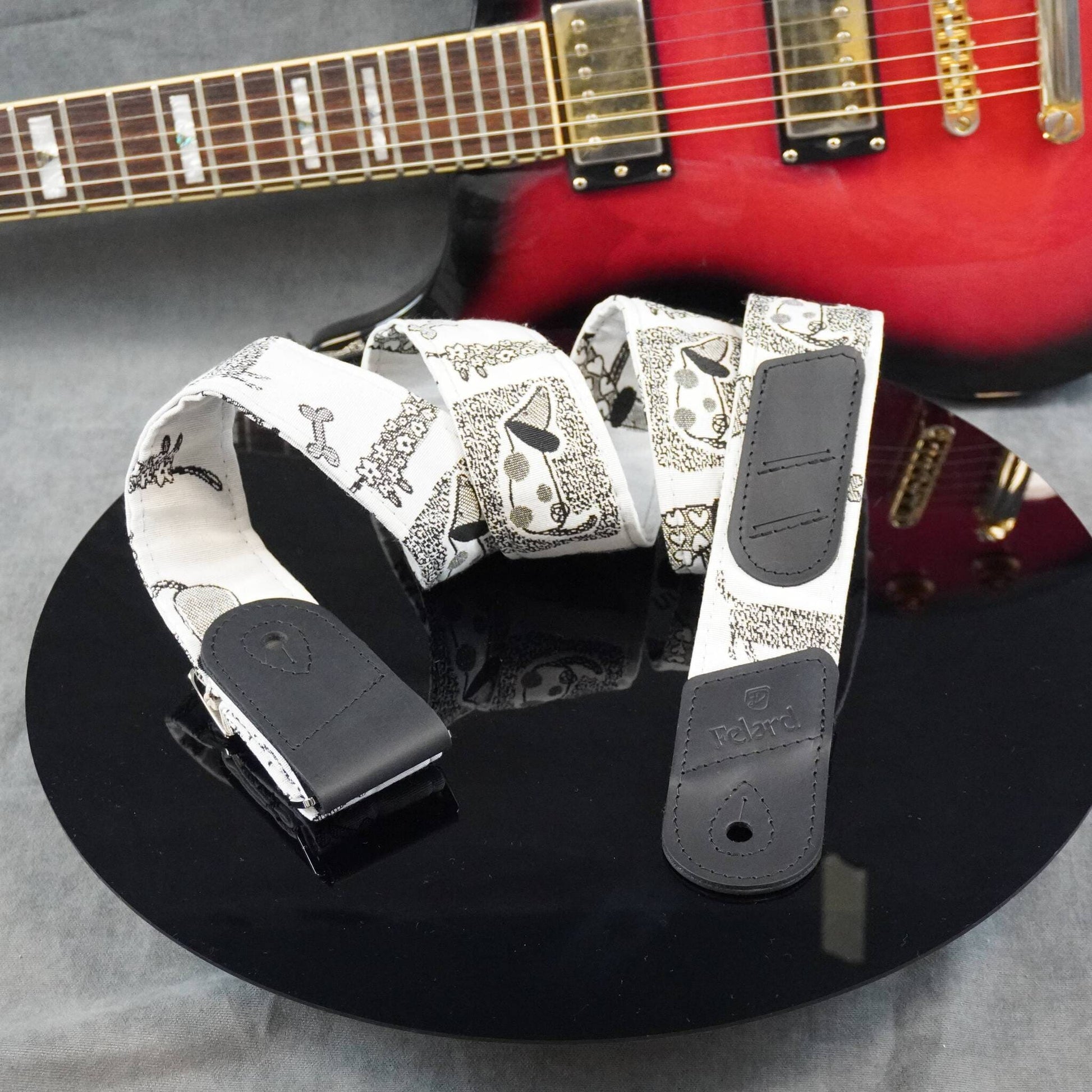 A white electric guitar strap with a black paisley pattern and black leather ends, displayed on a black reflective surface with a red electric guitar in the background.