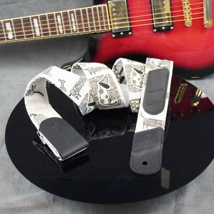A white electric guitar strap with a black paisley pattern and black leather ends, displayed on a black reflective surface with a red electric guitar in the background.