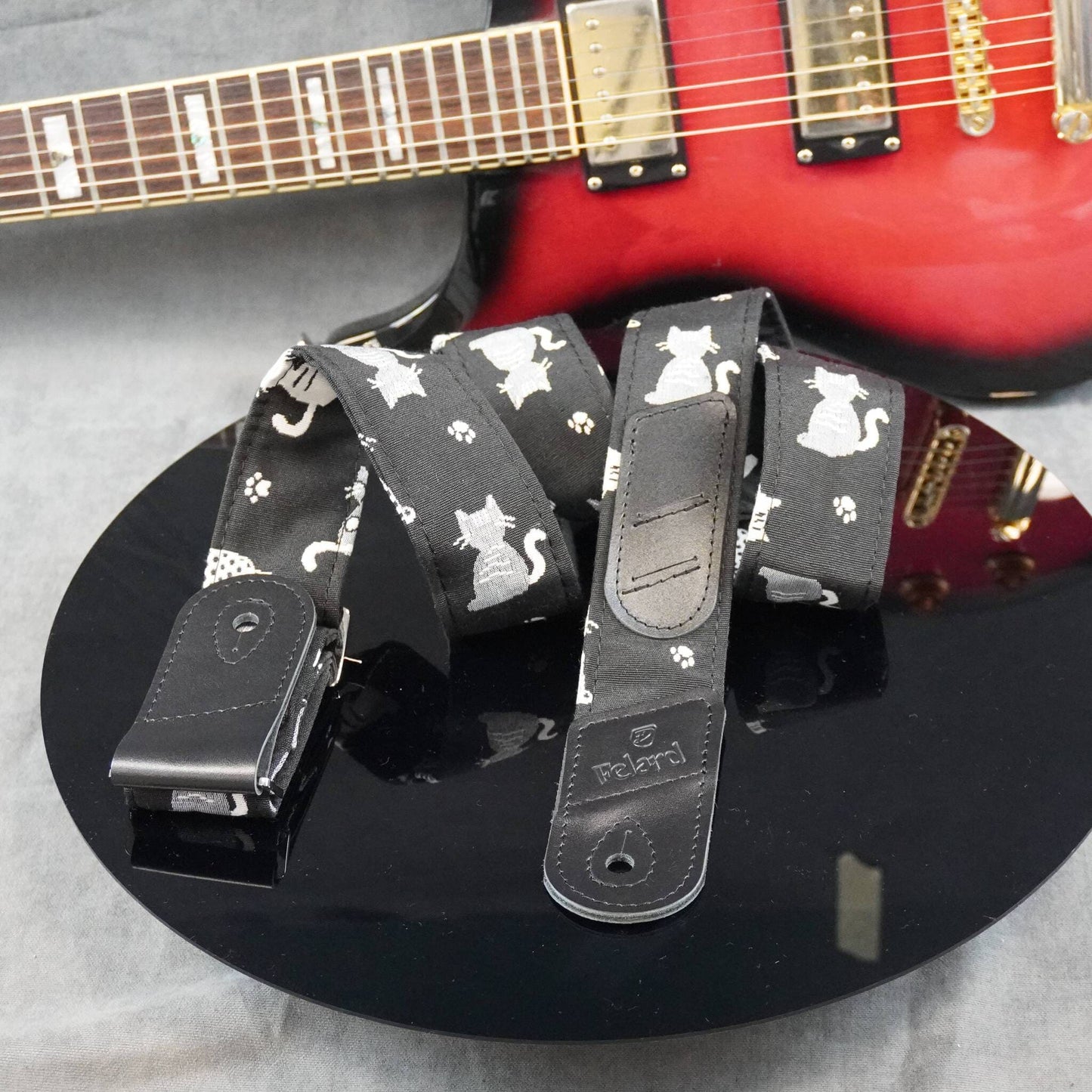 A black guitar strap with a white cat and paw print pattern, displayed on a black reflective surface with a red electric guitar in the background.