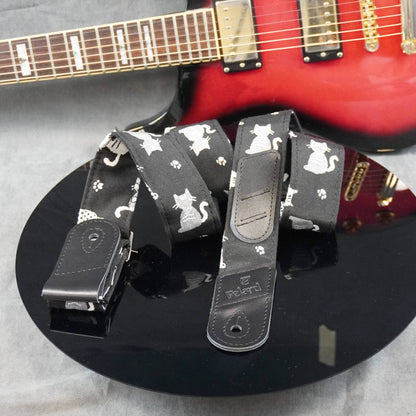 A black guitar strap with a white cat and paw print pattern, displayed on a black reflective surface with a red electric guitar in the background.