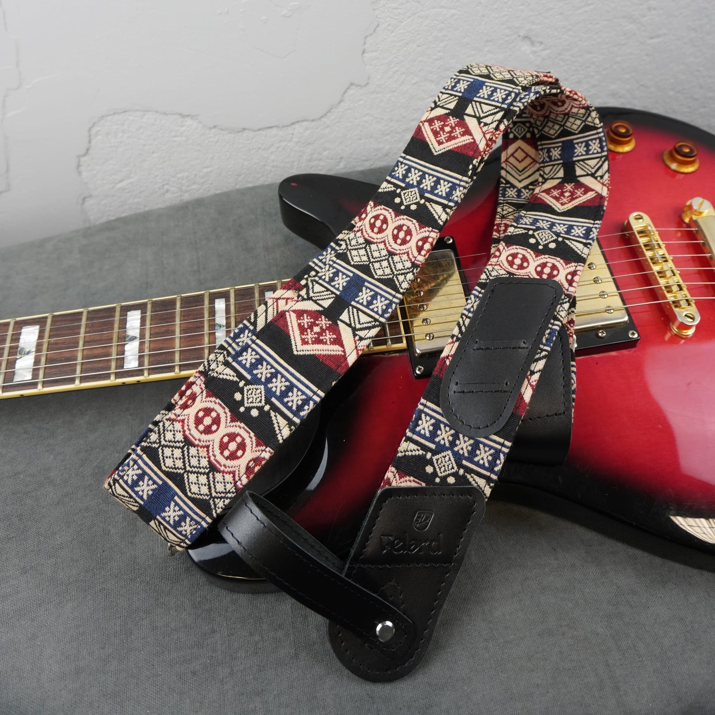 colorful geometric tribal pattern guitar strap in red, blue, beige, and black, draped over a red electric guitar on a gray surface.