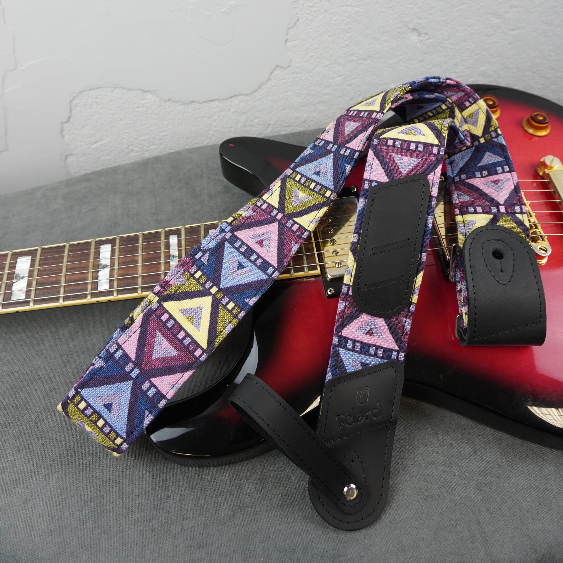 A guitar strap with a colorful geometric tribal pattern in purple, pink, blue, yellow, and white, laid over a red electric guitar on a gray surface.