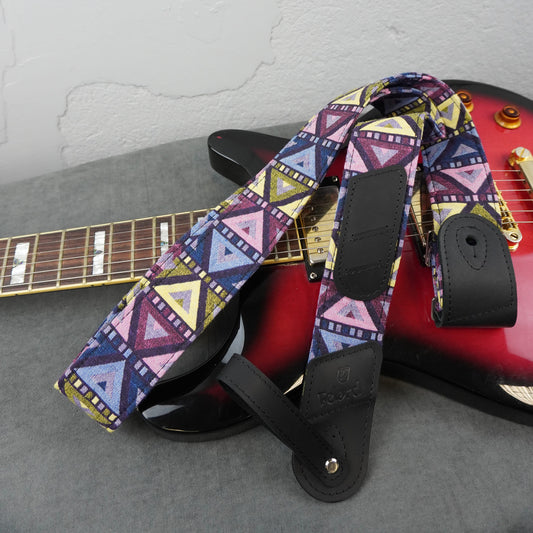 A guitar strap with a colorful geometric tribal pattern in purple, pink, blue, yellow, and white, laid over a red electric guitar on a gray surface.
