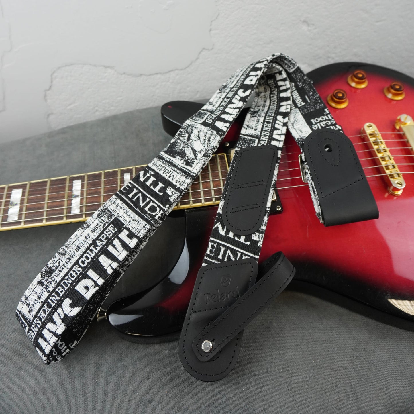 A guitar strap with a black and white newspaper print pattern and black leather ends, draped over a red electric guitar on a gray surface.
