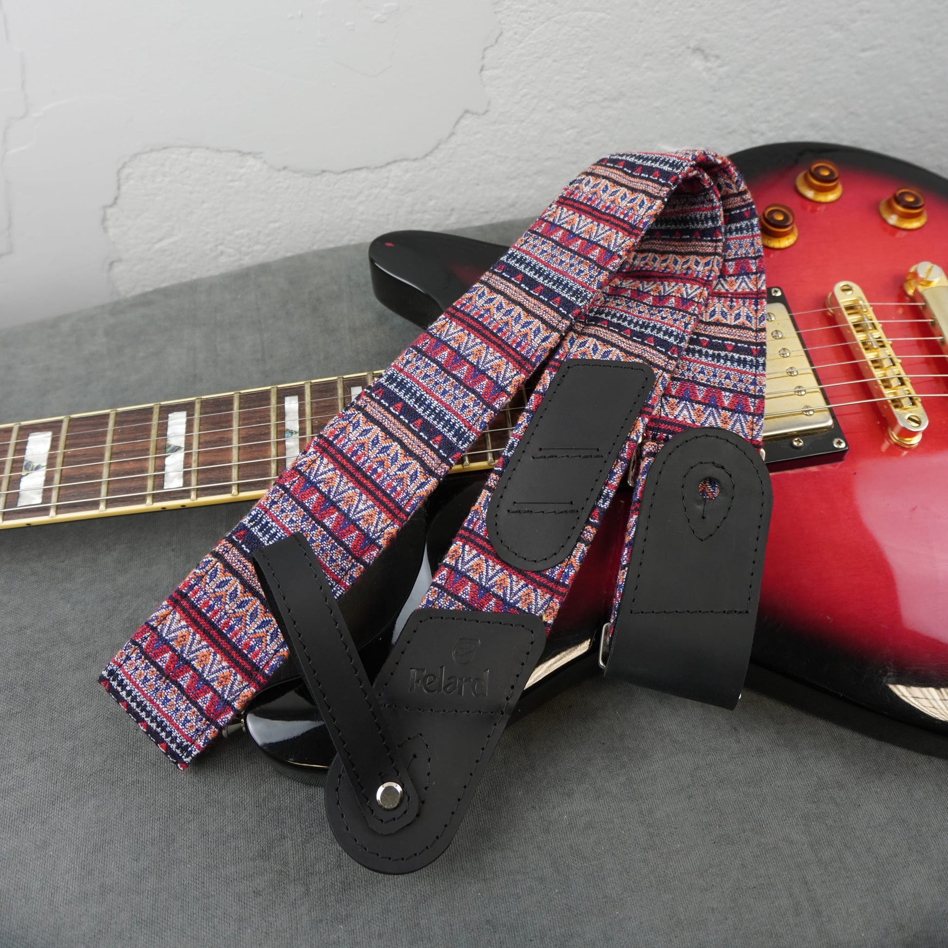 guitar strap with a colorful geometric tribal pattern in red, blue, and white, featuring black leather adjustable straps, displayed draped over a red electric guitar on a gray surface.