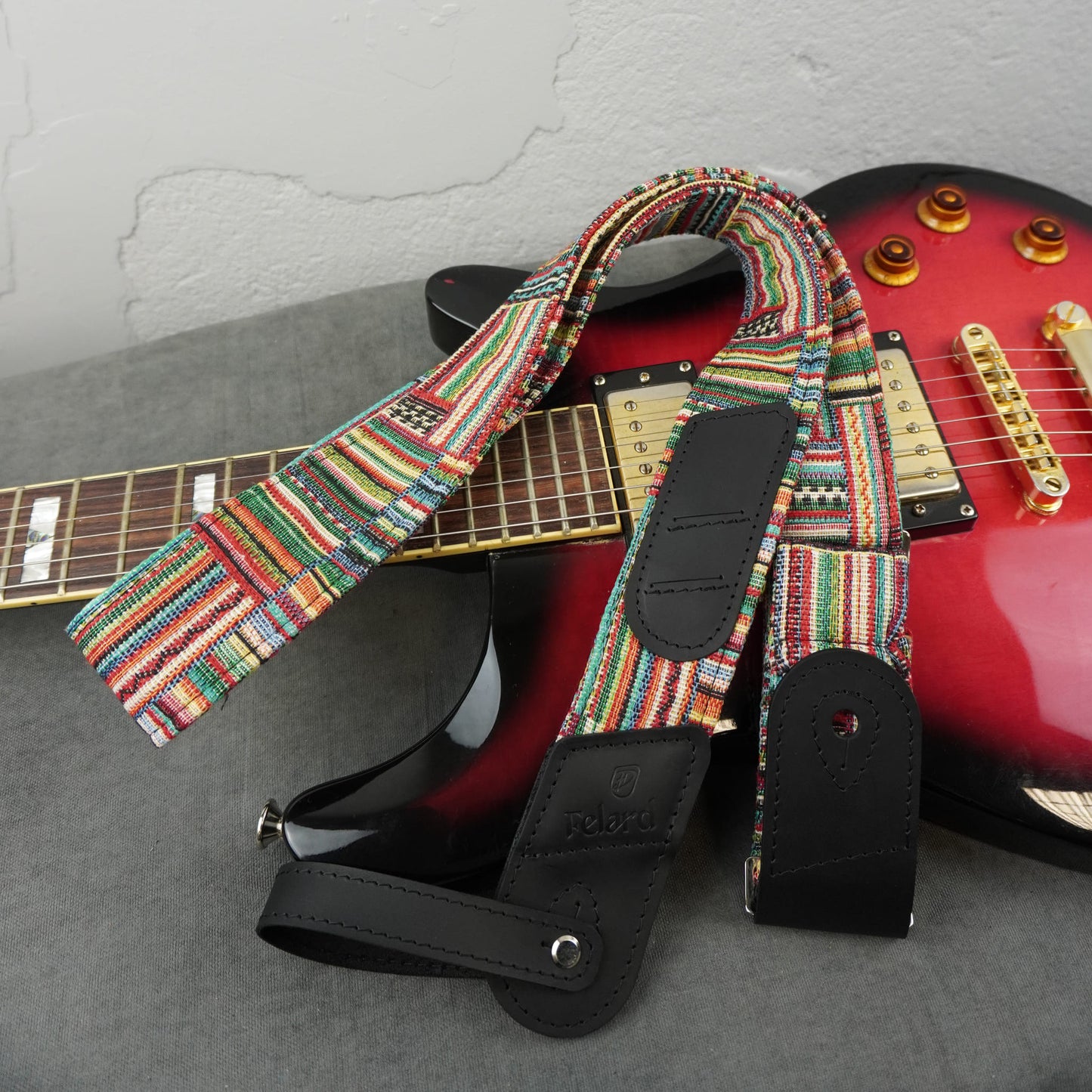A colorful woven guitar strap with black leather accents draped over a red electric guitar on a gray surface against a light wall.