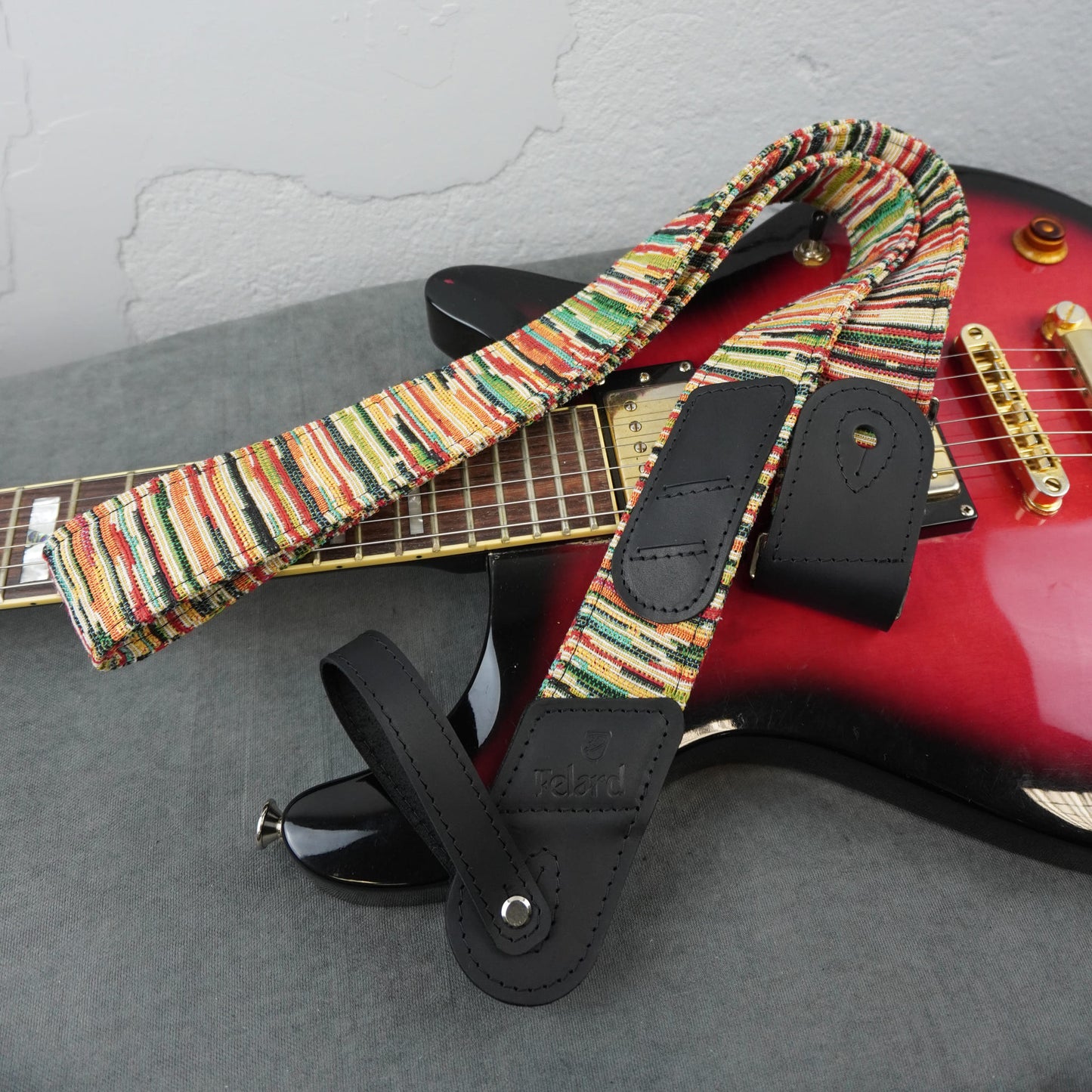Colorful striped pattern and black leather ends, draped over a red and black electric guitar on a gray surface.