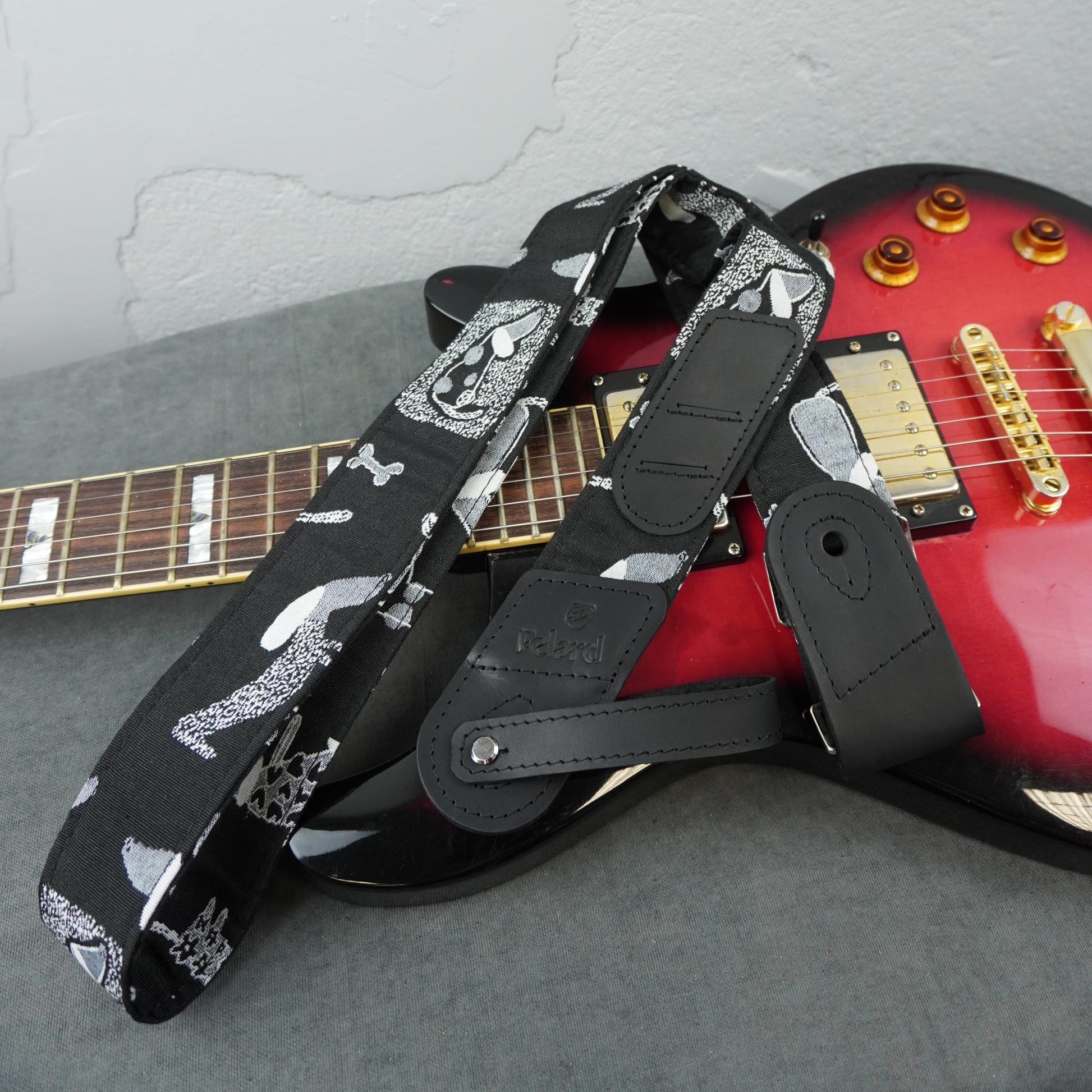 A black electric guitar strap with a white skull-and-crossbones pattern and black leather attachment sections, draped over a red electric guitar on a gray surface.