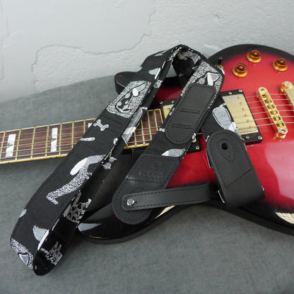 A black electric guitar strap with a white skull-and-crossbones pattern and black leather attachment sections, draped over a red electric guitar on a gray surface.