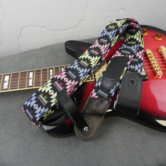 A guitar strap with a colorful geometric pattern in pink, yellow, green, black, and white, draped over a red electric guitar on a gray surface.