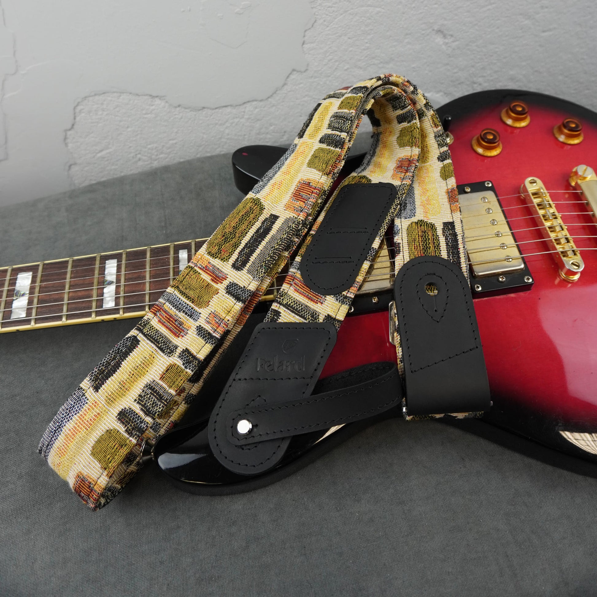 electric guitar strap with a multicolor geometric pattern in beige, brown, black, and red, featuring black faux-leather padding. The strap is draped over a red electric guitar on a gray surface against a textured gray wall.